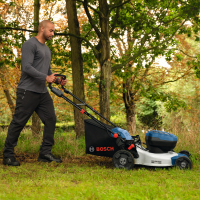 Homem a empurrar corta-relvas elétrico Bosch numa relva com árvores ao fundo.