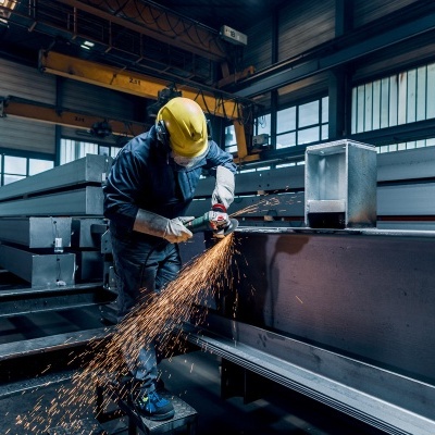 Trabalhador com capacete amarelo utilizando ferramenta elétrica em peça metálica cinzenta numa oficina industrial