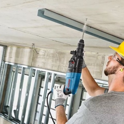 Homem com capacete amarelo a usar furadeira elétrica para perfurar teto de cimento no interior de construção