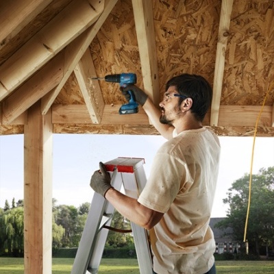Homem em escada a fixar madeira com furadeira azul numa estrutura de tecto em madeira
