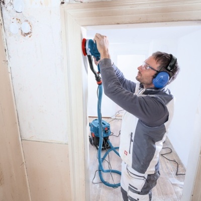 homem em roupa de trabalho usando lixadora elétrica azul numa porta de madeira