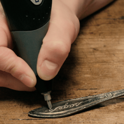 Pessoa usando ferramenta elétrica para gravar uma colher metálica sobre uma mesa de madeira.