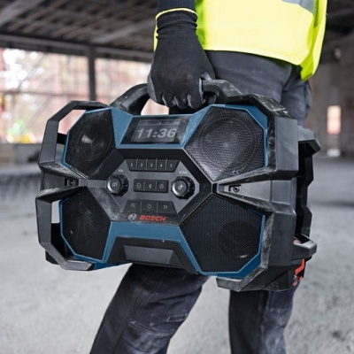 Rádio de construção Bosch preto e azul com visor digital e botões no centro, transportado numa obra.