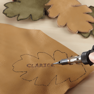 Gravação do nome 'CLARICE' em couro castanho claro com ferramenta de gravação, com folhas de couro verde e bege ao lado