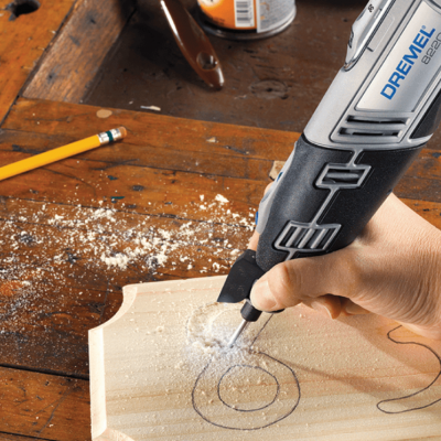 Pessoa usando ferramenta Dremel numa tábua de madeira com desenhos circulares num ambiente de trabalho em madeira.