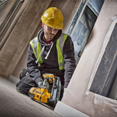 Trabalhador de construção com capacete amarelo e colete refletor utiliza ferramenta elétrica no chão da obra