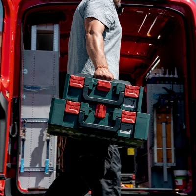 Mala de ferramentas verde com fechos vermelhos sendo transportada por pessoa junto a carrinha vermelha aberta