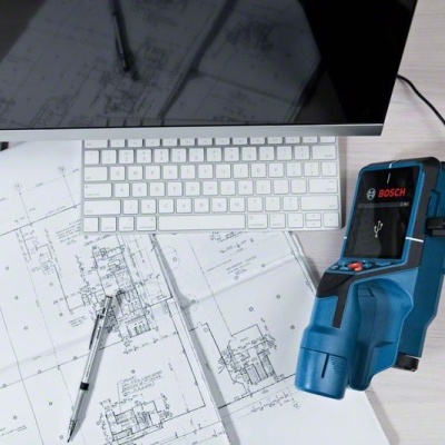 Estação de trabalho com teclado branco, monitor, desenhos técnicos, caneta preta e dispositivo BOSCH azul.