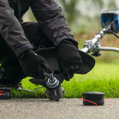 Pessoa ajusta lâmina de aparador Bosch preto e prateado junto a bateria EXPERT 18V preta e vermelha e carretel de linha em relvado.