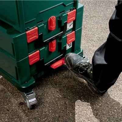 Caixa de ferramentas modular verde com fechos vermelhos e rodas, aberta com o pé por pessoa com calças pretas.