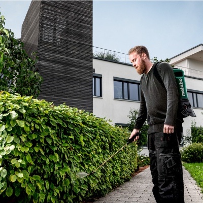 Homem a usar mochila pulverizadora verde para tratar relva junto de arbustos