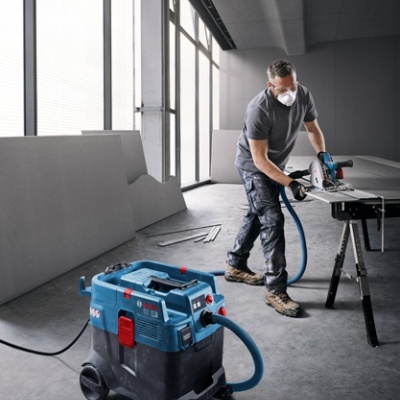homem operando serra circular Bosch azul ligada a aspirador industrial azul e preto numa oficina