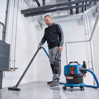 Homem usa aspirador industrial azul num ambiente interior com paredes brancas.