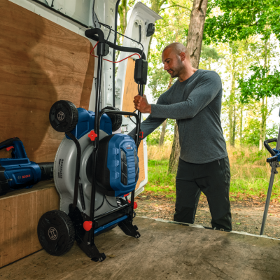 Homem manuseia cortador de relva elétrico azul e cinza dentro de van com paredes de madeira e árvores ao fundo.