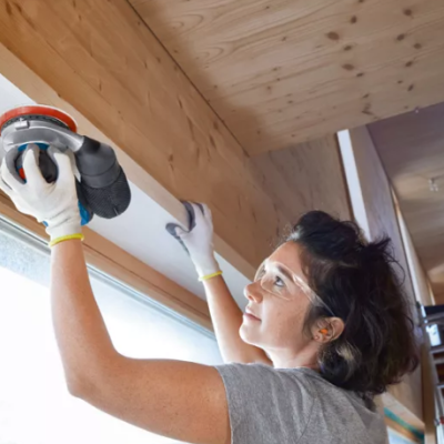 Mulher a lixar tábua de madeira clara no teto com lixadeira elétrica.