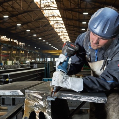 Homem com capacete e luvas a trabalhar metal numa oficina industrial.