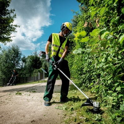 Homem corta relva com roçadora ao lado de sebes verdes num caminho de terra