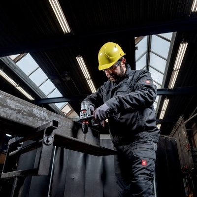 Homem com capacete amarelo e fato preto usa ferramenta elétrica em oficina industrial