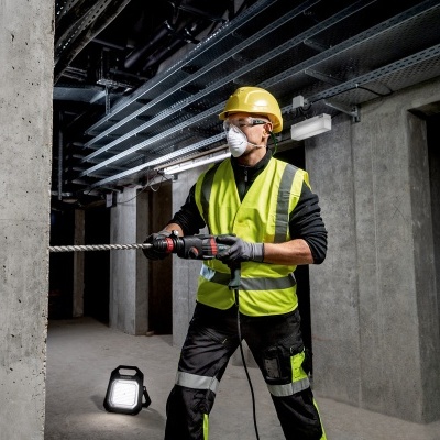 Trabalhador de construção com colete refletor, capacete amarelo e máscara operando ferramenta elétrica