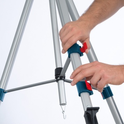 Tripé metálico prateado com detalhes azul e vermelho, mãos ajustando-o