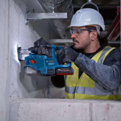 Homem com capacete branco e colete amarelo a usar furadeira Bosch azul em parede de cimento