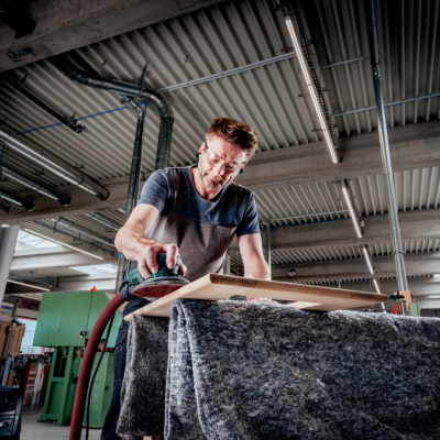 Homem a lixar uma tábua de madeira numa oficina industrial, com teto metálico e luz natural