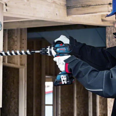 Trabalhador com capacete e luvas usa furadeira para perfurar madeira