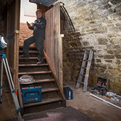 Escada de madeira e ferramentas Bosch numa cave com parede de pedra e homem na escada com telemóvel