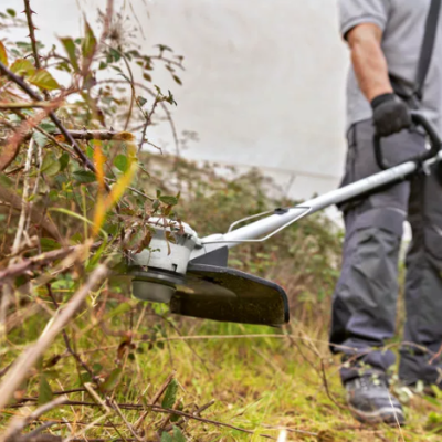 Aparelho cortador de relva cinza operado por pessoa com luvas e calças cinzentas em zona com plantas secas