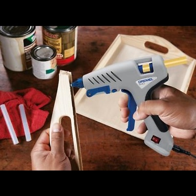 Pistola de cola quente DREMEL a ser usada para colar madeira, com latas de tinta e pano vermelho ao fundo.