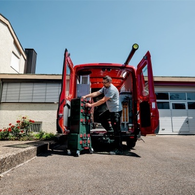Homem a manusear caixa verde com rodas na traseira de van vermelha aberta, em exterior junto a edifício branco com janelas.