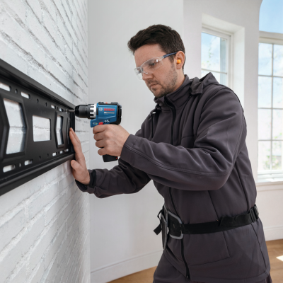 Homem usa furadeira Bosch para fixar estrutura numa parede de tijolo.