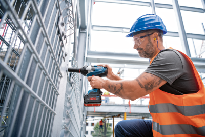 Trabalhador com colete e capacete a usar furadeira numa estrutura metálica num ambiente industrial