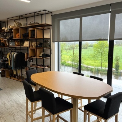 Mesa oval de madeira clara com cinco cadeiras altas pretas junto a janela e estantes com roupas e malas no interior