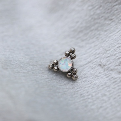 Piercing em aço inoxidável com pedra opala rodeada por esferas metálicas sobre superfície cinzenta