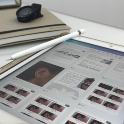 Tablet com caneta branca e fotografias de orelhas na tela, livro e relógio ao fundo
