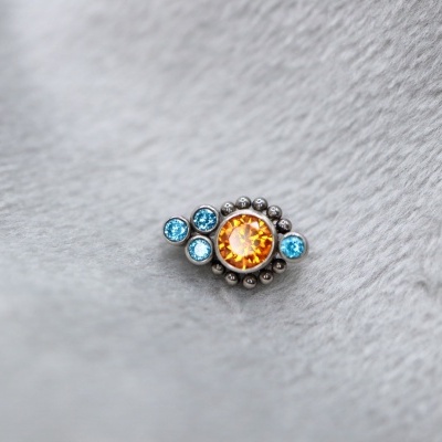 Anel com pedra laranja central e pedras azuis sobre fundo cinza
