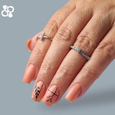 Mão feminina com unhas decoradas em pêssego e desenhos pretos, usando anéis prateados