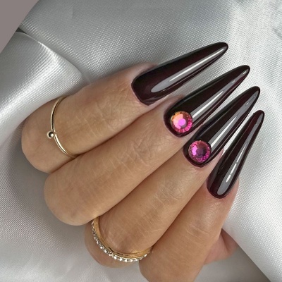 Unhas castanho escuro brilhantes com pedras rosa e anéis dourados em mão sobre tecido prateado