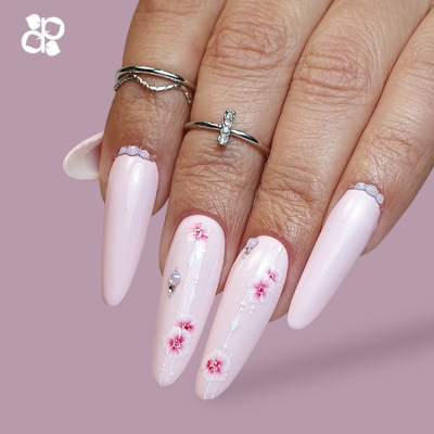 Mãos com unhas longas rosa claro decoradas com flores e anéis prateados.