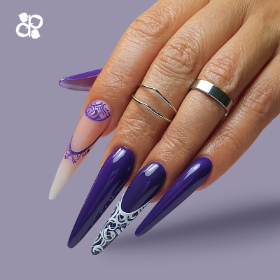 Mão com unhas stiletto roxas e desenhos, anéis prateados, fundo cinzento lilás.