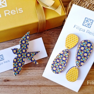 Brincos e broche coloridos com padrões geométricos em cartões da marca Filipa Reis junto a uma caixa amarela com laço.