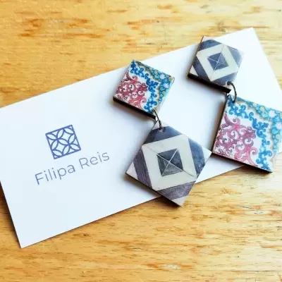 Brincos geométricos e florais de Filipa Reis sobre cartão branco em cadeira de madeira.