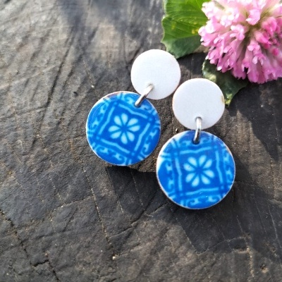 Brincos redondos branco e azul com padrão floral sobre madeira escura e flor cor-de-rosa