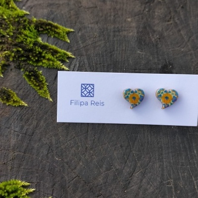Brincos em forma de coração com padrão floral amarelo e azul num cartão branco com o nome Filipa Reis sobre uma superfície de madeira escura com musgo