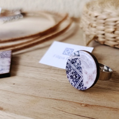 Anel com placa redonda decorada em padrão azul e branco, em superfície de madeira com cartões da marca
