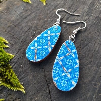 Brincos ovais azuis com padrão branco e linhas douradas em fundo de madeira com musgo