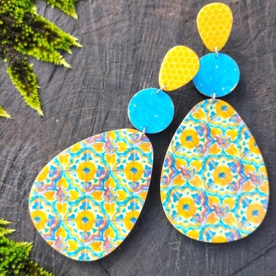 Brincos coloridos com padrão floral sobre madeira escura e folhagem verde