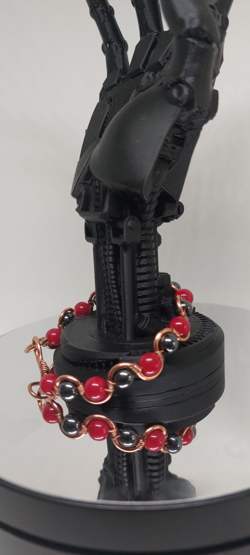 Pulseira com bolas vermelhas e cinzentas em fio de cobre numa mão robótica negra.