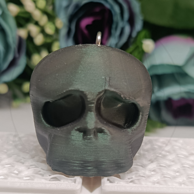 Pendente caveira preto em plástico com textura sobre base branca com flores ao fundo